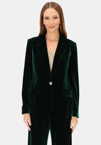 Zielony aksamitny blezer w kolorze szmaragdowym z klapą z wcięciem, pojedynczym zapięciem na guzik, dwoma przednimi kieszeniami i dekoracyjnym akcentem w postaci pszczoły.