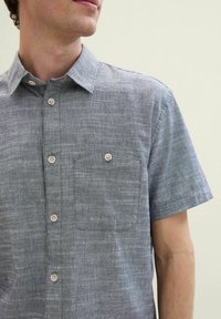 Cămașă cu mânecă scurtă și nasturi din țesătură gri deschis, având un buzunar pe piept, guler rotund și nasturi bej. Design texturat, casual.