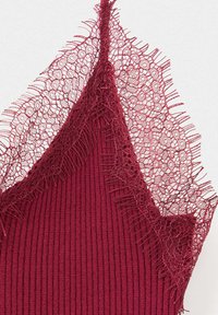 Gros plan d'un tissu côtelé bordeaux avec une délicate bordure en dentelle florale et une fine bretelle sur fond blanc.