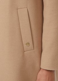 Manteau en laine beige avec une poche latérale et un bouton circulaire doré. Le tissu a une texture douce et une finition soignée et ajustée.