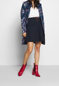 Laivansininen luisteluhame, jossa on nauhakuja ja taskut, yhdistettynä kuviolliseen pitkään takkiin, jossa on sinisiä, punaisia ja valkoisia kuvioita. Punaiset korkeat nilkkabootsit.