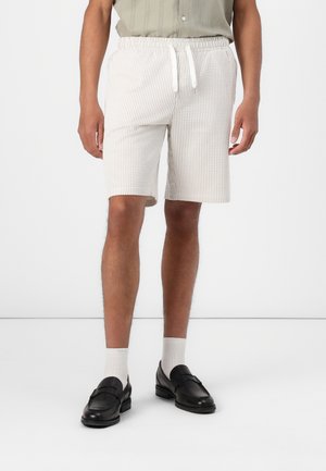 Shorts à rayures blanches et beige avec une taille élastique et un cordon de serrage. Coupe ajustée, longueur genoux, associés à des mocassins noirs et des chaussettes blanches.