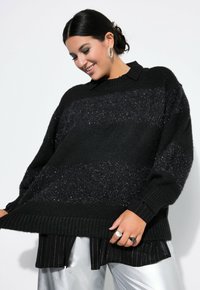 Pullover in maglia nera oversize con strisce orizzontali testurizzate, polsini e orlo a coste, indossato sopra una camicia a righe in bianco e nero. Visibili dettagli in argento.