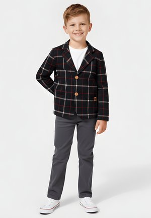 Uśmiechnięty młody chłopiec w czarnej, kraciastej marynarce, białej koszuli, szarych spodniach i białych trampkach stoi z jedną ręką za plecami.