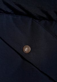 Merikankaan kangas, jonka pinta on sileä ja jossa on pyöreä pronssinappula, jossa on karhun muotoilu, ommeltuna kankaan taitokseen.