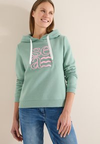 Sweat à capuche vert menthe avec un motif texte graphique rose. Comprend une capuche à cordon, des poignets et un ourlet côtelés, associé à un jean bleu.