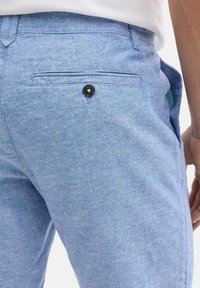 Pantalones de algodón azul claro que muestran un bolsillo trasero con un botón oscuro. Tejido ligeramente texturizado. Bolsillo lateral visible.