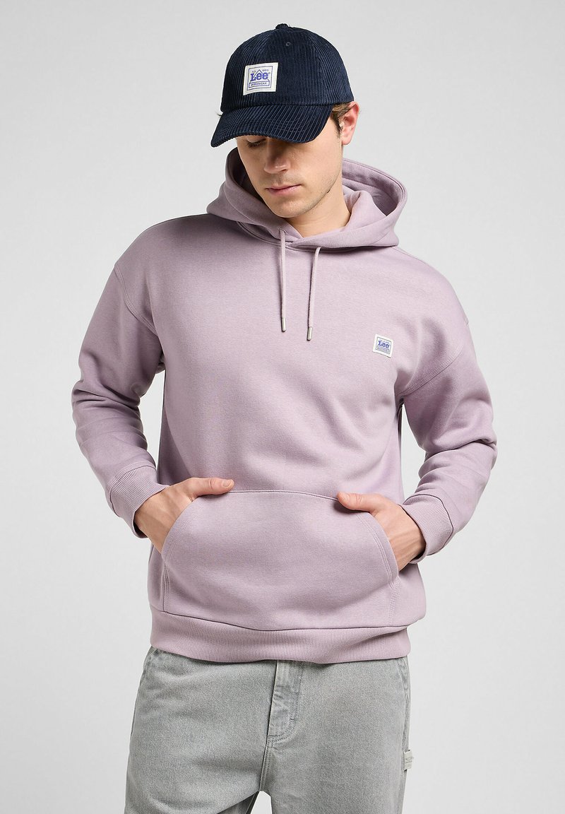 Light lavender hoodie with a kangaroo pocket, ribbed cuffs, and a small logo patch. Paired with a dark navy corduroy cap.