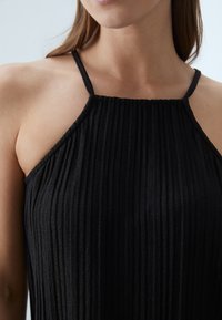 Haut plissé noir avec des bretelles fines, un décolleté droit et un tissu texturé. Le design met en avant des lignes verticales.