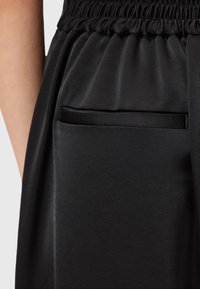 Zwarte elastieken broek met een close-up van een achterzak en een gedeeltelijk zicht op de arm van een persoon.