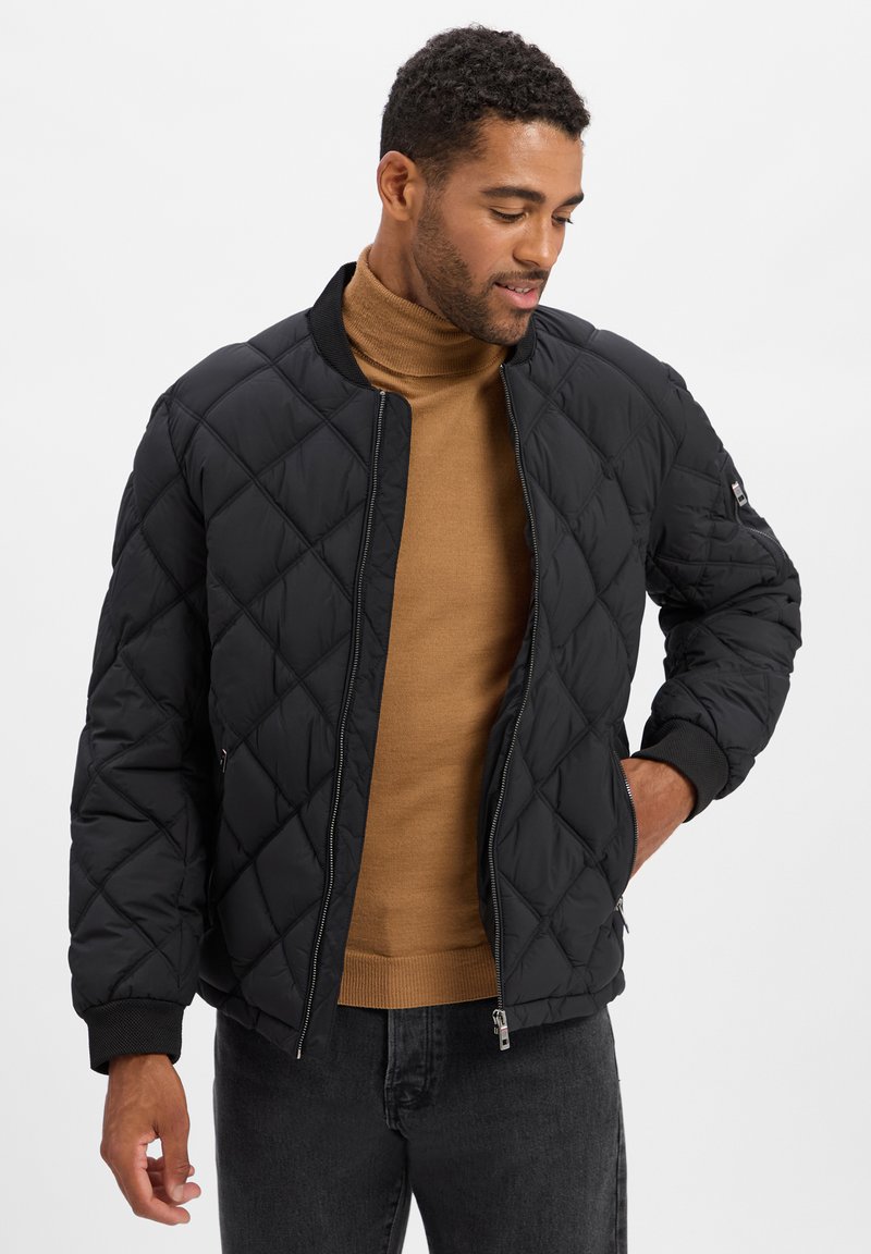 Homme portant une veste bomber matelassée noire sur un pull à col roulé marron et un jeans noir, avec une main dans la poche de la veste.