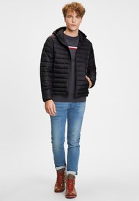 Svart vadderad jacka med huva, med quiltad struktur och dragkedja fram. Matchas med en grå tröja, ljusa jeans och bruna snörskor.