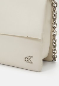 Borsa in pelle beige con patta pieghevole, dotata di una tracolla in catena argentata e un discreto logo "CK" impresso sul davanti.