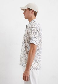 Korte mouw shirt in lichtbeige met zwart bloemenpatroon. De stof lijkt lichtgewicht. Gecombineerd met een bijpassende lichte pet.