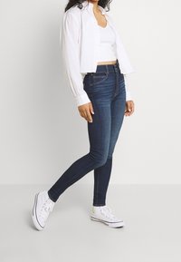 Slim jeans i mörkblå denim, tillsammans med en vit crop top och en vit skjorta med knappar. Vita sneakers fulländar outfiten.