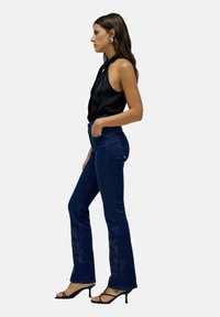 Haut halter en satin noir avec un décolleté noué, associé à un jean évasé bleu foncé orné de broderies florales près des ourlets et des talons fins noirs.