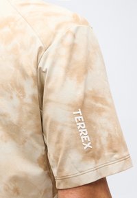 Detailní záběr na béžové a světle hnědé tričko s krátkým rukávem a efektem batiky, na rukávu je vertikálně vytištěný nápis "TERREX".