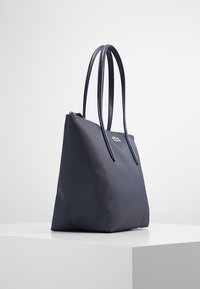 Navy tote bag in textured synthetic material, featuring two long handles and a zip closure, with a small green logo accent on the front.