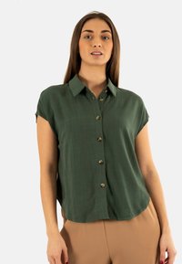 Blusa verde a maniche corte con frontale con bottoni, realizzata in tessuto leggero. Presenta un colletto classico e una texture sottile.