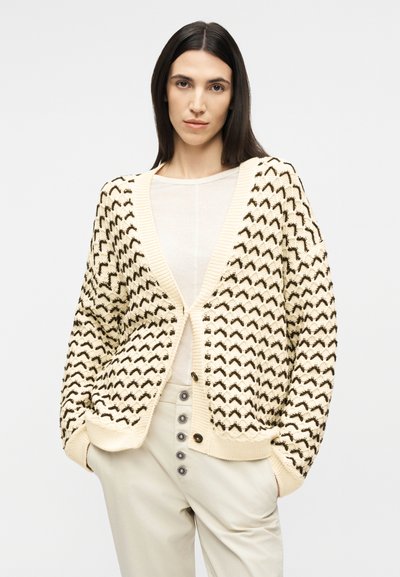 Γυναίκα με ίσια σκούρα μαλλιά φοράει κρεμ ζακέτα με μαύρο σχέδιο σε σχήμα κεβρόν, λευκό τοπ και μπεζ παντελόνι με ψηλή μέση και διακοσμητικά κουμπιά.