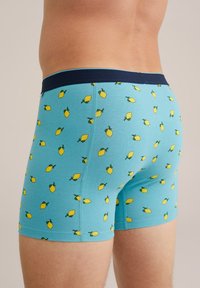 Plave bokserice s uzorkom limuna, s tamnoplavim elastičnim pojasom i glatkom teksturom. Skupljaju tkaninu oko bedara.