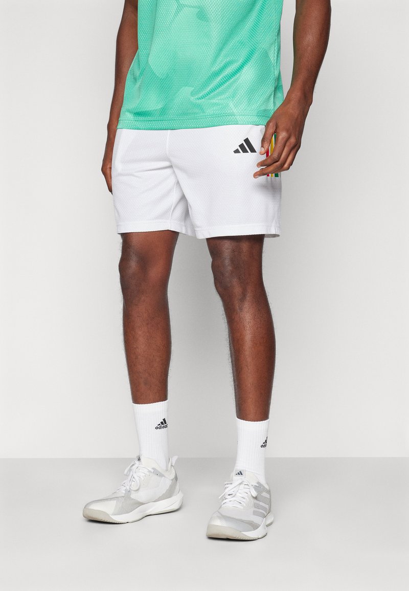 Calções atleticos brancos com um design texturizado, combinados com sapatos cinzentos e meias brancas, apresentando logótipos da Adidas em preto. T-shirt de malha verde.
