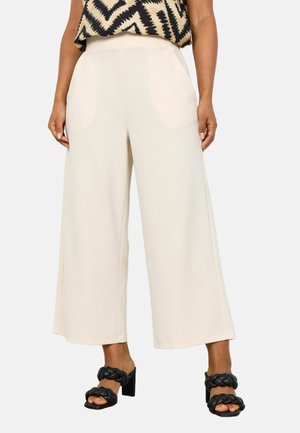 Beige wide-leg pants made of soft material with side pockets, paired with black heeled sandals featuring a braided strap design.