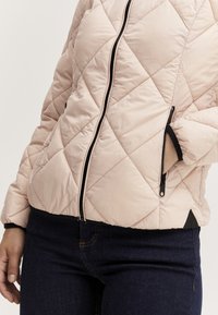 Donna che indossa una giacca trapuntata rosa chiaro con dettagli neri della cerniera e jeans blu scuro, con una mano nella tasca della giacca.