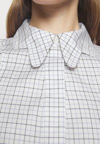 Camisa de vestir con cuello, de tejido blanco con patrones a cuadros en azul y negro. Cierre de botones en el cuello. Textura ligera.