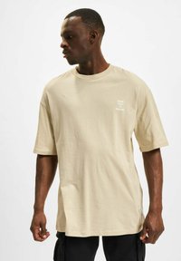 Beige bomulls t-skjorte med korte ermer, avslappet passform, sidesplitter og trykt tekst på venstre bryst, kombinert med svarte cargobukser.