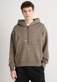 Sudadera color caqui con cremallera frontal, que presenta bordados coloridos de animales, un bolsillo tipo canguro y puños acanalados en las mangas y el dobladillo.