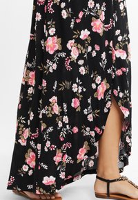 Rochie maxi florală din material negru, cu flori roz și albe, cu fanta pe lateral și sandale maro cu bretele decorative.