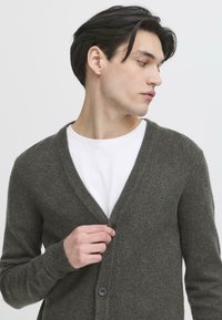 Grå ullcardigan med v-hals, knappeåpning og ribbede mansjetter, lagd over en hvit t-skjorte. Glatt tekstur, minimalistisk design.