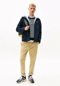 Chaqueta ligera de marinero con capucha, forro de felpa blanca, suéter a rayas, pantalones caqui y zapatillas azules con detalles blancos. Mochila colgada de un hombro.