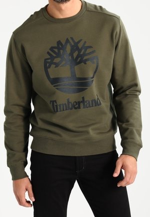 Mies, joka on pukeutunut oliivinvihreään Timberland-collegepaitaan, jossa on musta puun logo, ja mustiin housuihin, yksinkertaista taustaa vasten.