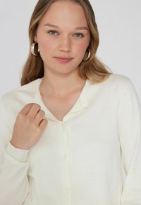 Gebreide cardigan in zachte crème kleur, met een afgeronde kraag, knoopsluiting en geribbelde manchetten. Model houdt lichtjes het gebied van de kraag vast.