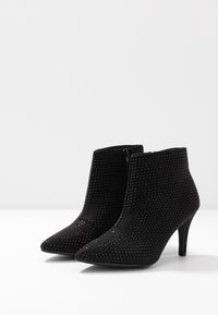 Botines de ante negros con punteras afiladas, detalles de tachuelas, cremalleras laterales y tacones altos y delgados sobre una superficie blanca.