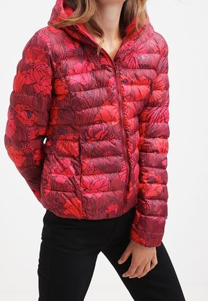 Femme portant une doudoune rouge à motif floral avec capuche, fermée, associée à un pantalon noir, debout avec une main près de la hanche.