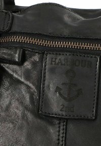 Section d'un sac en cuir noir montrant une fermeture éclair en laiton et un patch cousu avec un symbole d'ancre et le texte "HARBOUR 2nd".