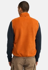 Fleece vest in bright orange with a high collar, featuring a navy long-sleeve base, ribbed cuffs, and a smooth texture.