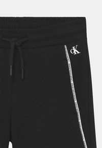Černé tepláky s žebrovaným pasem, šňůrkou a bílým logem. Obsahují boční pruh s nápisem "CALVIN KLEIN JEANS".