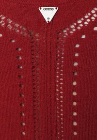Červený pletený svetr s otevřeným švem a otvory, s trojúhelníkovou stříbrnou visačkou s logem "GUESS" na výstřihu. Texturovaný povrch.
