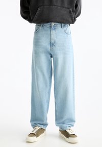 Jeans de pierna ancha de color azul claro, hechos de denim, con una superficie texturizada y diseño clásico de cinco bolsillos, combinados con zapatillas marrones y blancas.