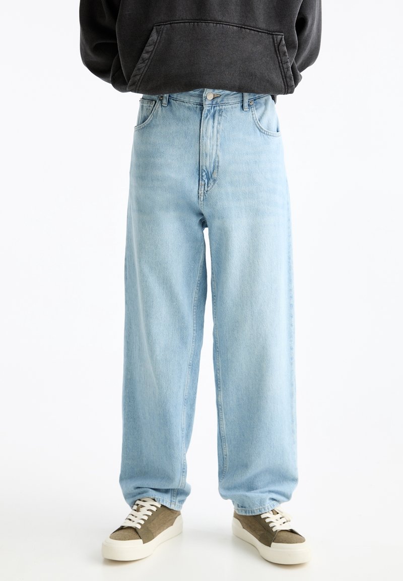 Jeans de pierna ancha de color azul claro, hechos de denim, con una superficie texturizada y diseño clásico de cinco bolsillos, combinados con zapatillas marrones y blancas.