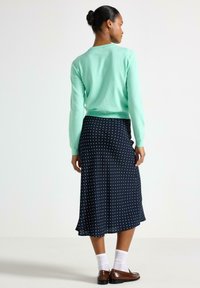 Maglione verde chiaro a maniche lunghe abbinato a una gonna midi blu navy a pois. La gonna ha un orlo svolazzante. Scarpe marroni e calzini bianchi completano l'outfit.