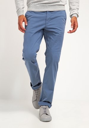 Mężczyzna idzie, ubrany w jasnoniebieskie spodnie, szare tenisówki i jasnoszarą koszulę z długim rękawem na białym tle.