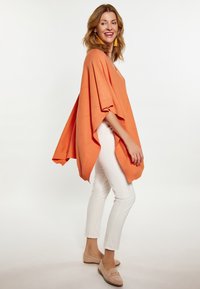 Orange strikket poncho med løs pasform, der har hulmønstre, korte ærmer og ribkant. Sammen med hvide slim-fit bukser og beige sko.