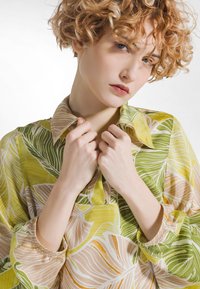 Blomstret bluse i grøn og gul med krave, lange ærmer med rynkede manchetter og en let, glat tekstur.