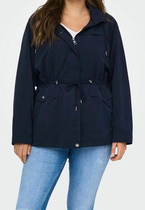 Femme portant une veste zippée bleu marine avec taille à cordon et poches à rabat devant, associée à un jean bleu clair.