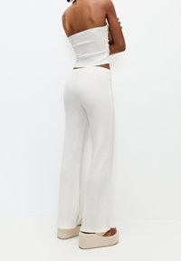 Femme portant un haut tube en maille blanche et un pantalon large assorti, pieds nus sur des sandales plateforme beiges, contre un fond blanc.